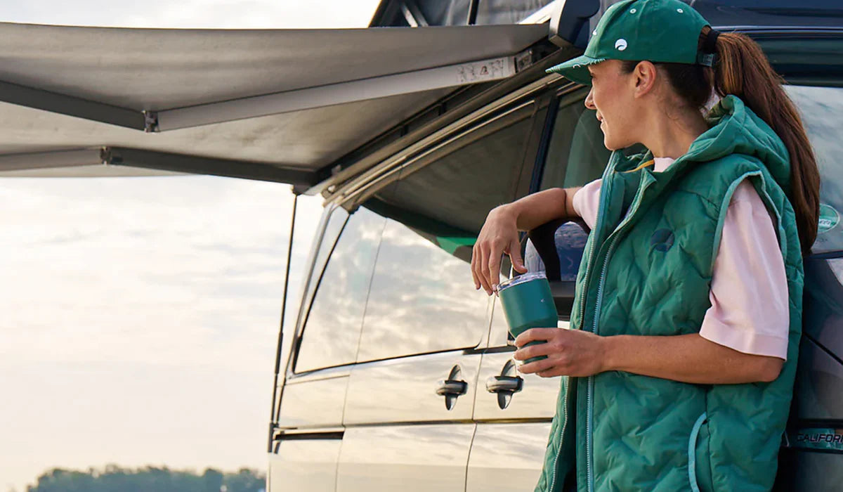 Frau mit Thermobecher neben Campervan unter ausgefahrener Markise beim Camping
